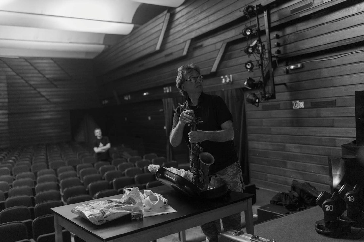 In this black and white image of a small theater with wood paneling and lighting instruments, a man wearing glasses, camo pants, and a dark t-shirt assembles his saxophone on stage. The theater is mostly empty except for one blurred man standing in the seats half way to the back watching the person on stage.