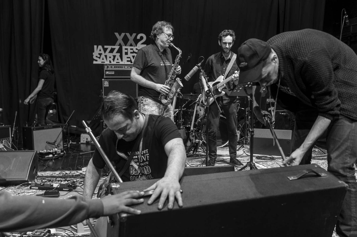 In this black and white soundcheck image, two musicians play on stage full of microphones and other musical equipment. The man playing sax on the left wears glasses, light camo pants, and a dark t-shirt. The man playing guitar wears all black and has dark hair and beard. Three technicians are also visible adjusting and moving gear on the stage.