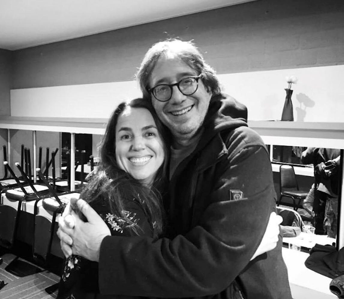 In this black and white image a woman with long brown straight hair wears a dark top with light stitched flowers and hugs a man wearing glasses and a dark hoodie. Both are looking at the camera. They look to be in a theater greenroom.