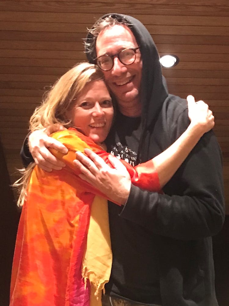 A blonde woman wearing a bright orange top and yellow scarf hugs a man wearing glasses and a black hoodie, both are looking at the camera.