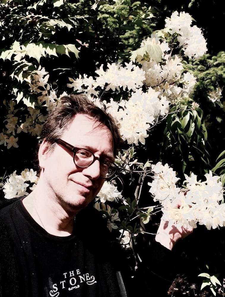 A middle-aged white man wearing glasses, a hoodie, and a black t-shirt that reads The Stone stands in front of a tall bush with white flowers.