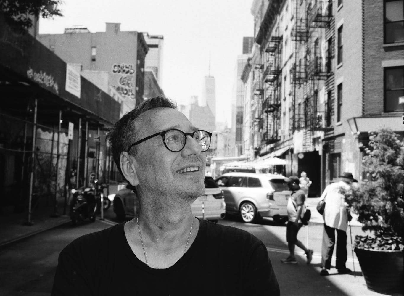 A black and white image of a white man wearing a black t-shirt and glasses standing on a street in NYC. Buildings both low and high in the background as well as two passersby.