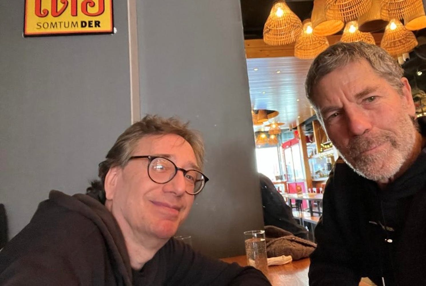 Two middle aged white men with graying hair look at the camera in a cafe. John Zorn is wearing glasses and is wearing a dark hoodie. Ohad has a slight grey beard and is wearing a dark shirt.