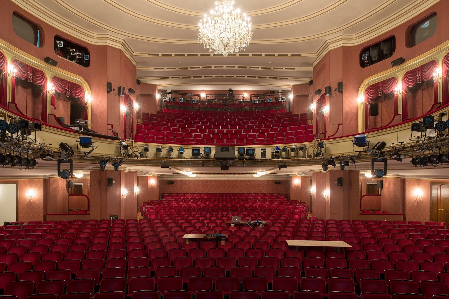 Interior of a large theater house with a chandiler, red seats, and gold trim.