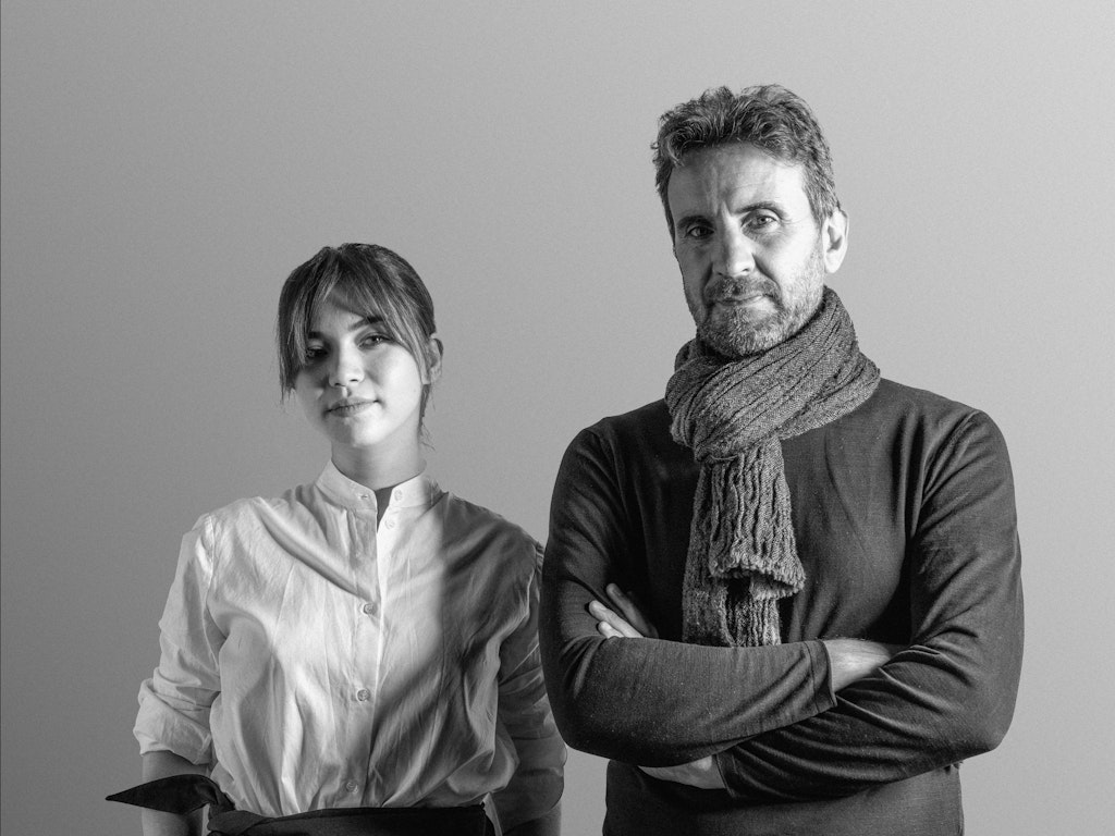 A black-and-white image of a woman in a white shirt and a man in a scarf stand side by side against a plain background.