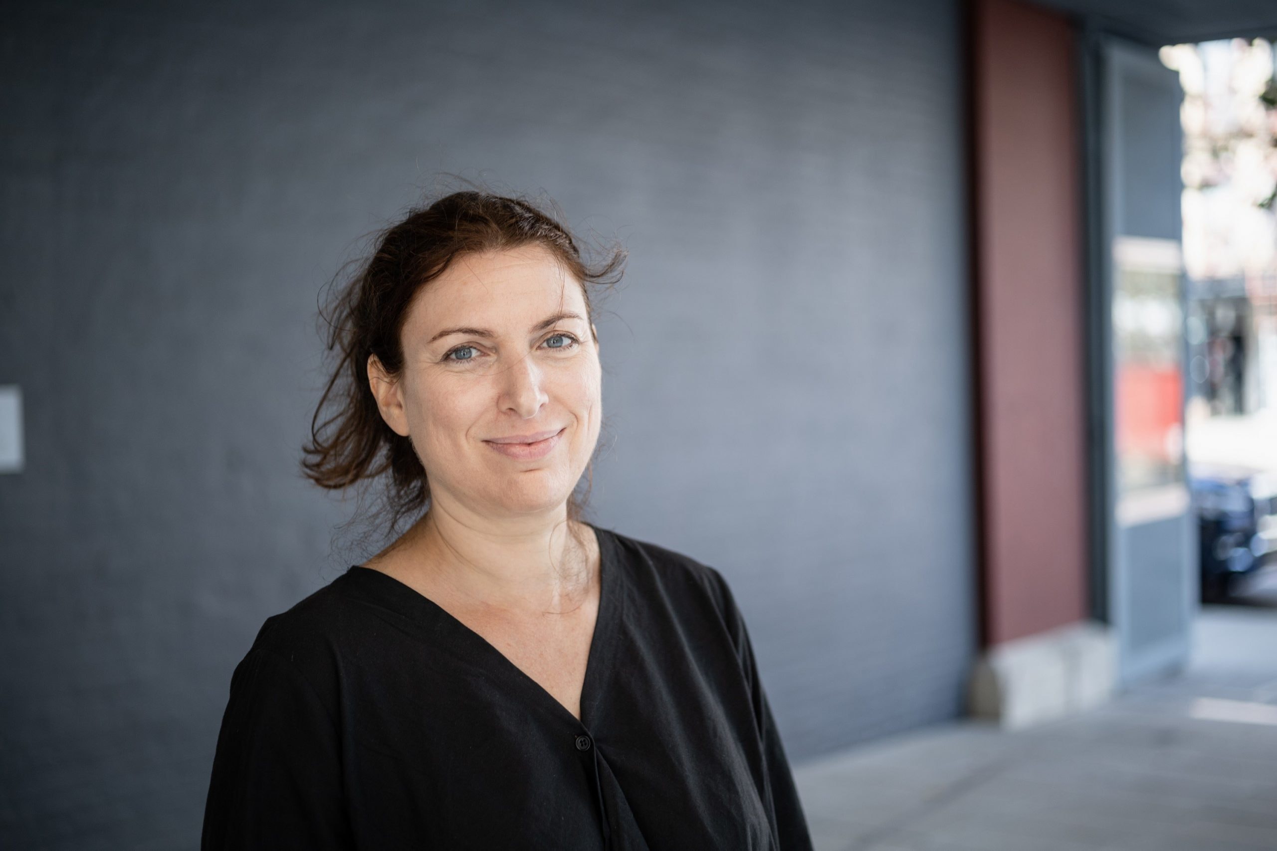 A white woman in a black shirt stands before a dark gray wall.