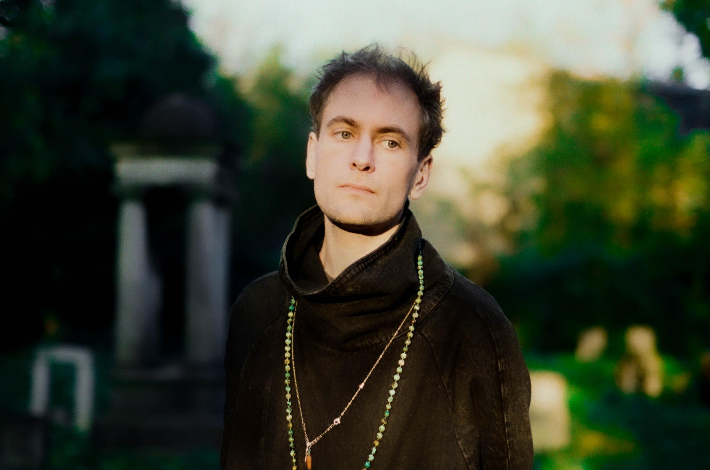 A person with light skin and short brown hair is standing outdoors, gazing to the right, and wearing a black shirt with two long necklaces.