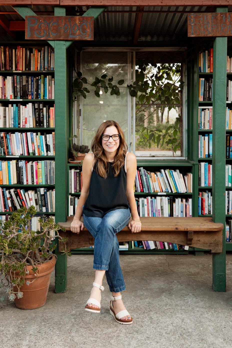 A woman wearing glasses, a sleeveless black top, and jeans sits outside on a bench near potted plants, in front of green bookshelves filled with colorful books.