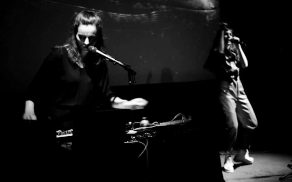 A shadowy, black-and-white image of two people on a small stage. Both stand and sing into mics. The person on the left plays electronic instruments on a table in front of them.