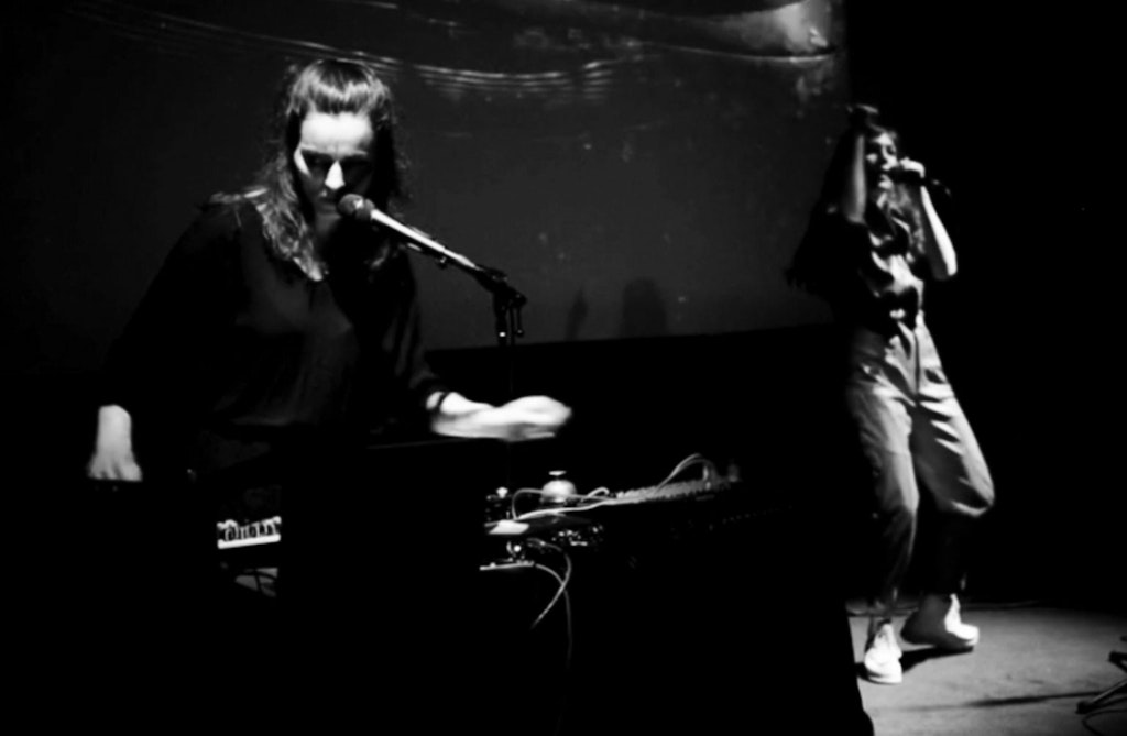 A shadowy, black-and-white image of two people on a small stage. Both stand and sing into mics. The person on the left plays electronic instruments on a table in front of them.
