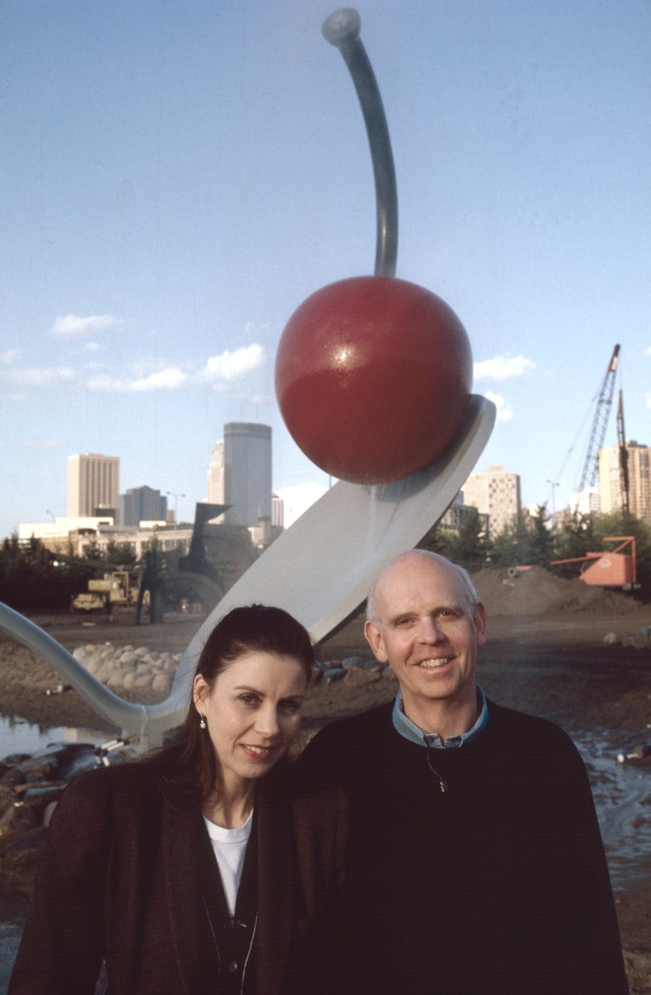 A man and a woman stand in front of a large public sculpture of a cherry sitting on the tip of a spoon.