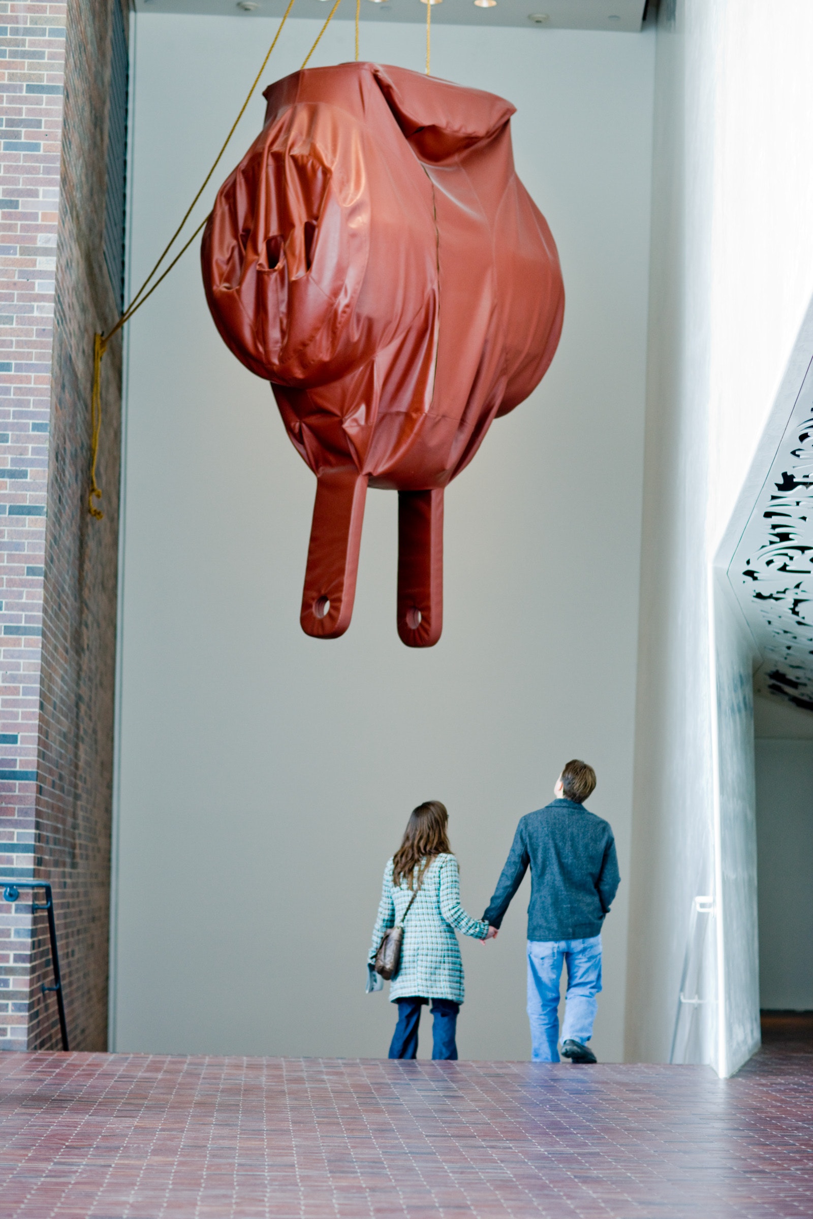 A couple holds hands while looking at a large soft sculpture of a three way electrical plug in a museum stairwell.