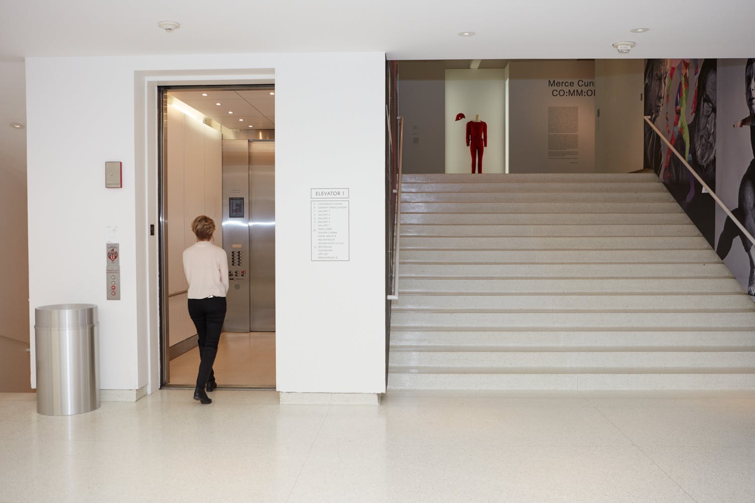 An adult weaing a white top and black pants enters an elevator that is located next to stairs leading up to an art gallery.