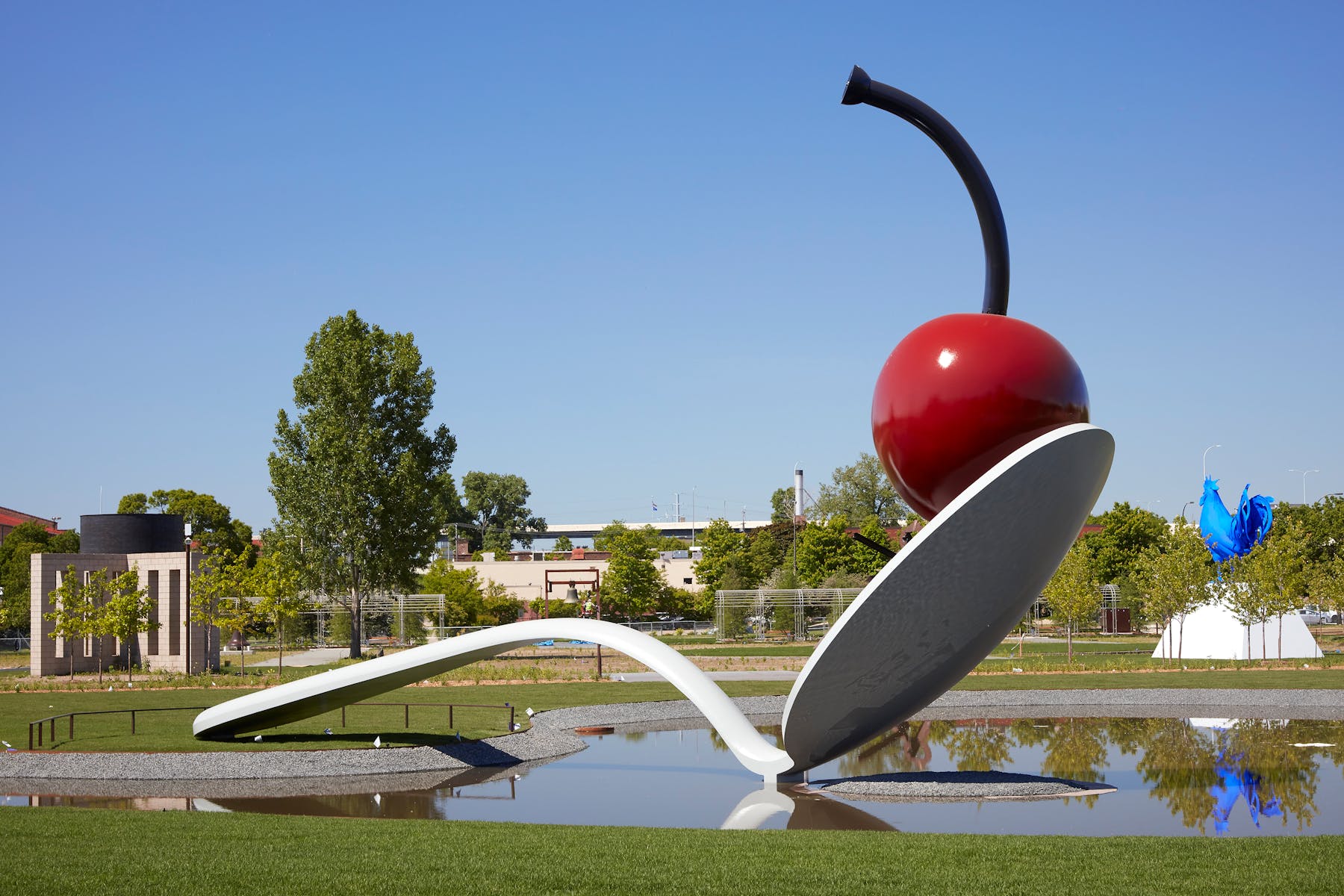 Large sculpture of red cherry balancing on tip of white spoon, above small pond, with sculptures and trees in the background
