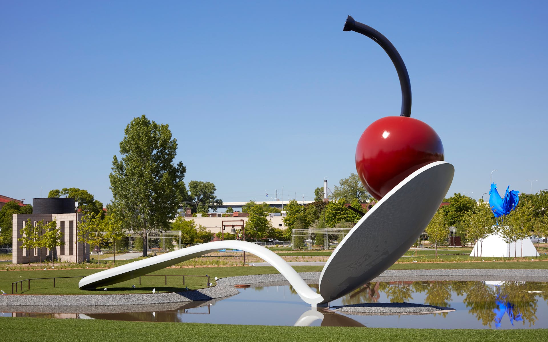 Image of large spoon and cherry sculpture on top of pond, with trees and sky in the background