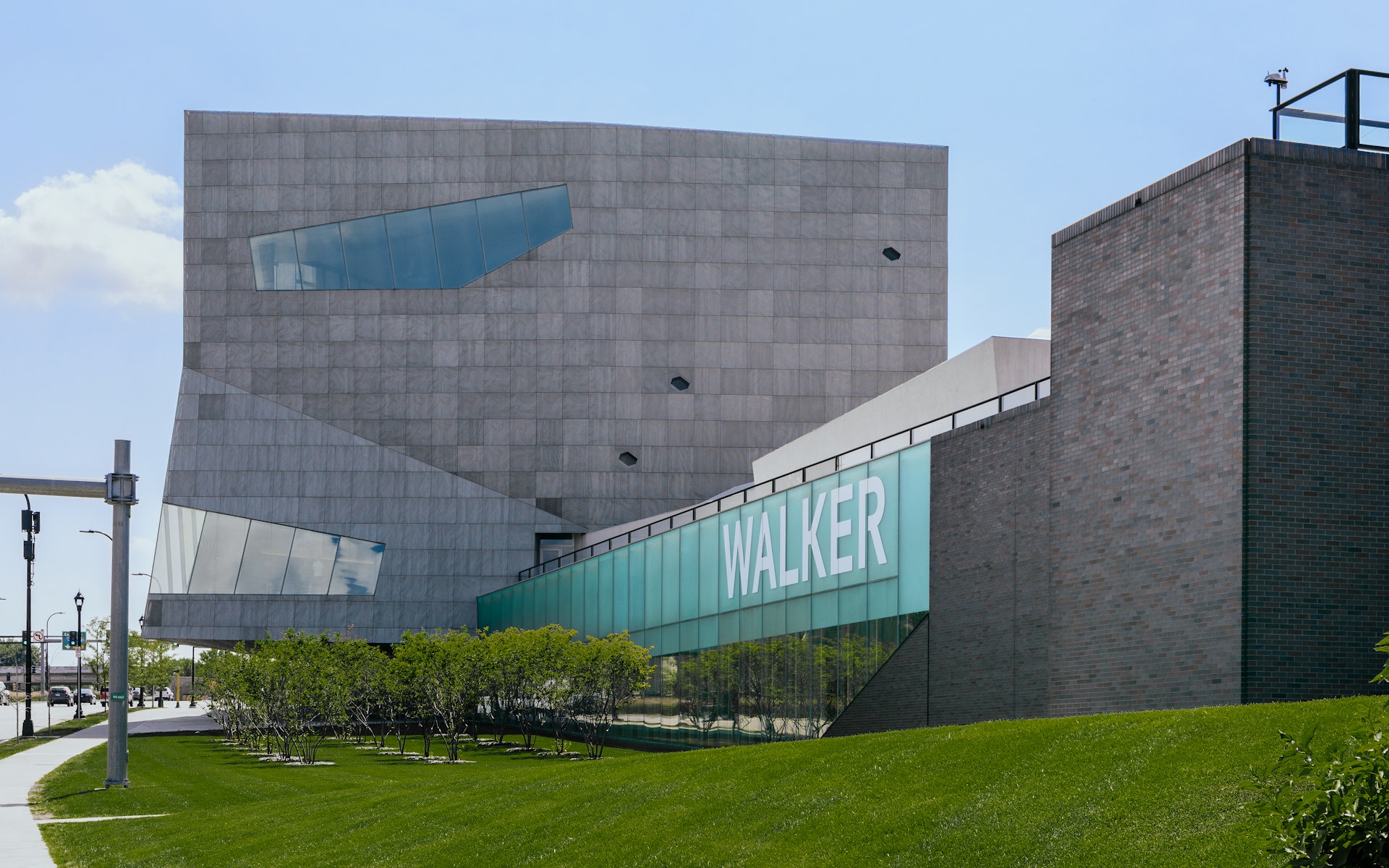 View of the Walker Art Center Campus from Hennepin Avenue, including the Herzog and de Meuron expansion theater bloc and a partial view of the brick Barnes Buidling at right. Also shown, small grove of trees outside Hennepin Lounge, and large WALKER letters on glass wall.