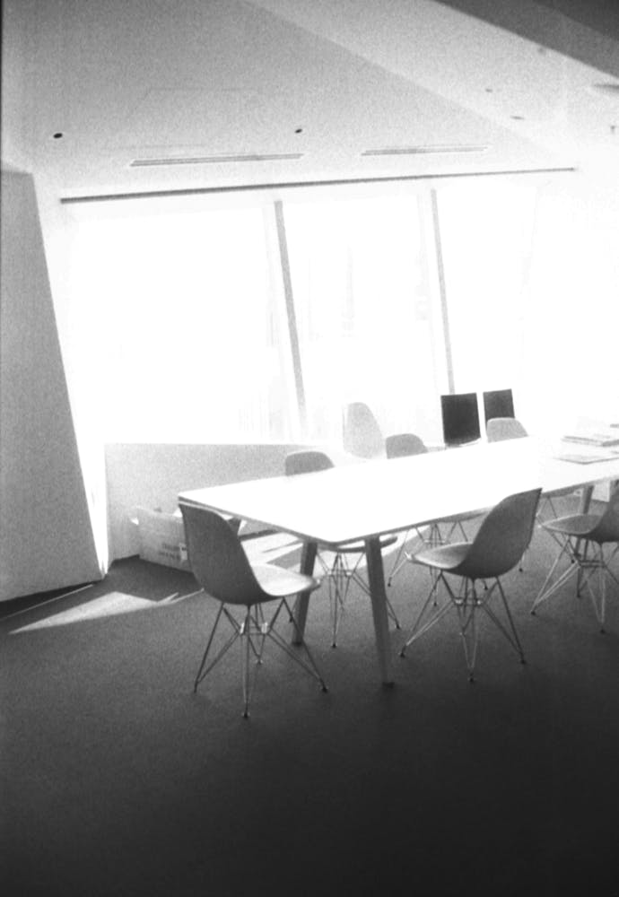 Black and white image of an empty meeting table and chairs next to a large angular window.