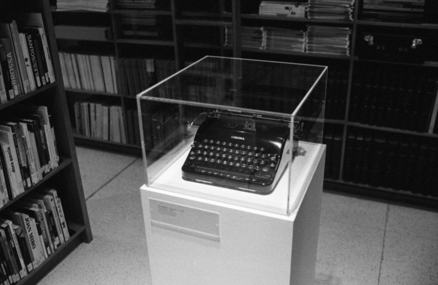 A typewriter sints under a plexiglass box on a pedestal among rows of books in a library.