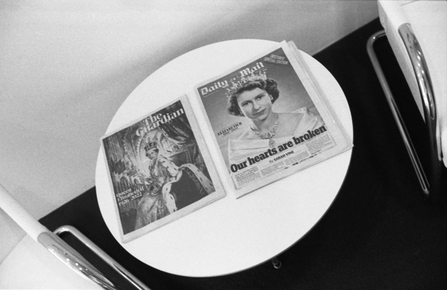 Two newspapers with headlines about the death of Queen Elizabeth II sit on modern end table in between two chairs in a waaiting room.