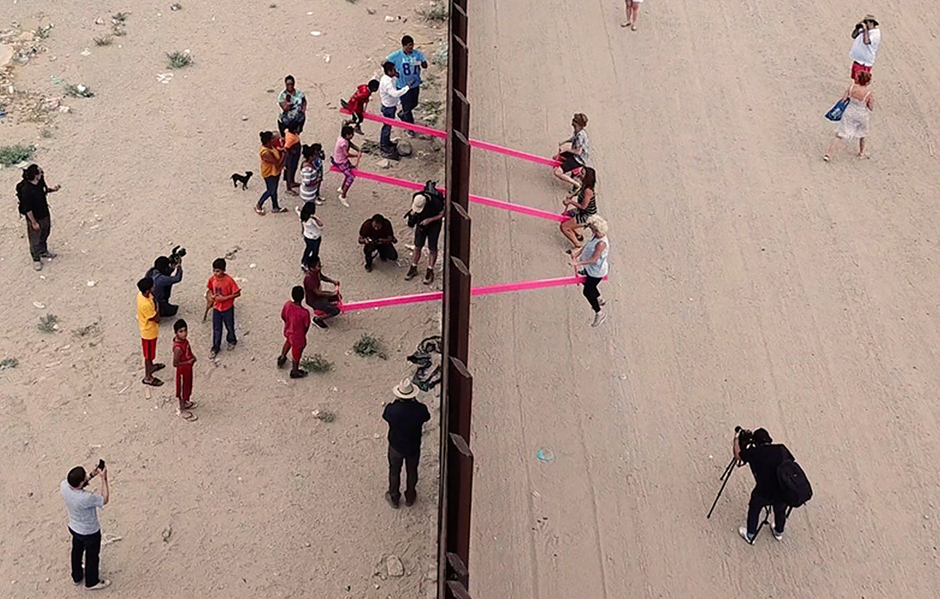 Image of children playing on pink seesaws on either side of a border wall
