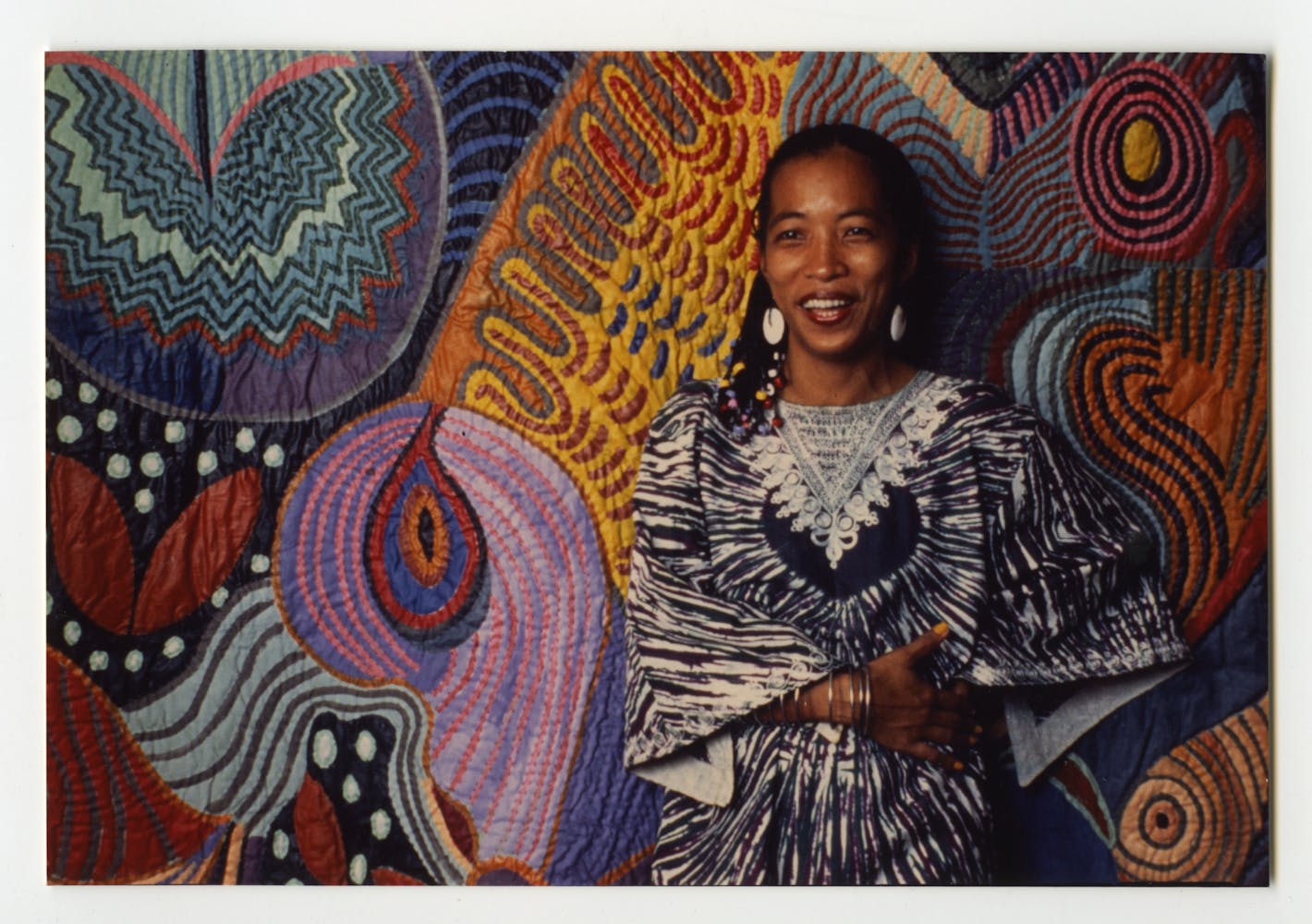A woman in a bright dress smiles while standing in front of a colorful painting.
