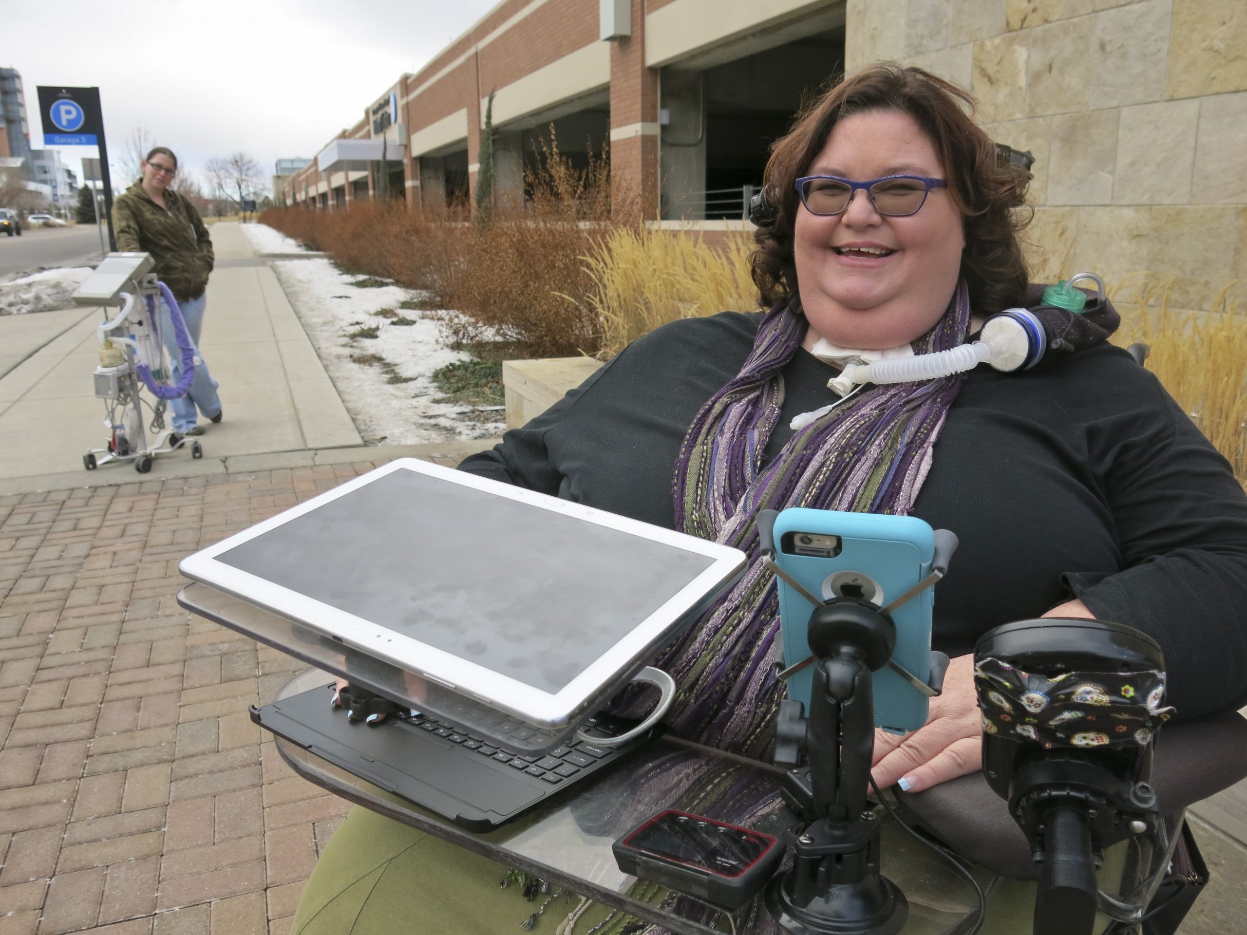A white wheelchair user with a tracheostomy and tinted glasses smiles on the street. Another person stands in the background with a medical device on wheels.