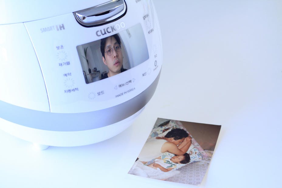 A medium-skinned performer's face is reflected in the screen of a rice cooker. A printed photo of two figures sleeping sits on the table nearby.