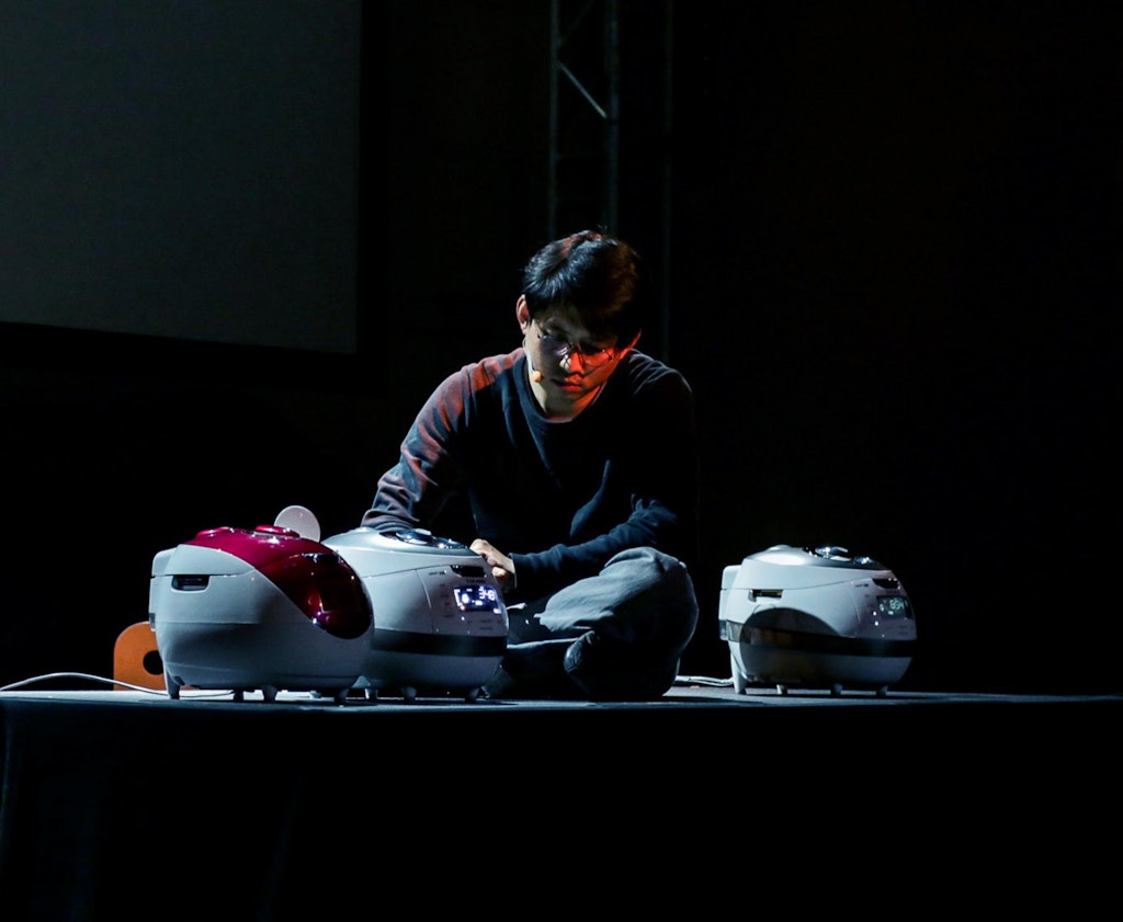 A performer with medium skin sits next to three rice cookers.
