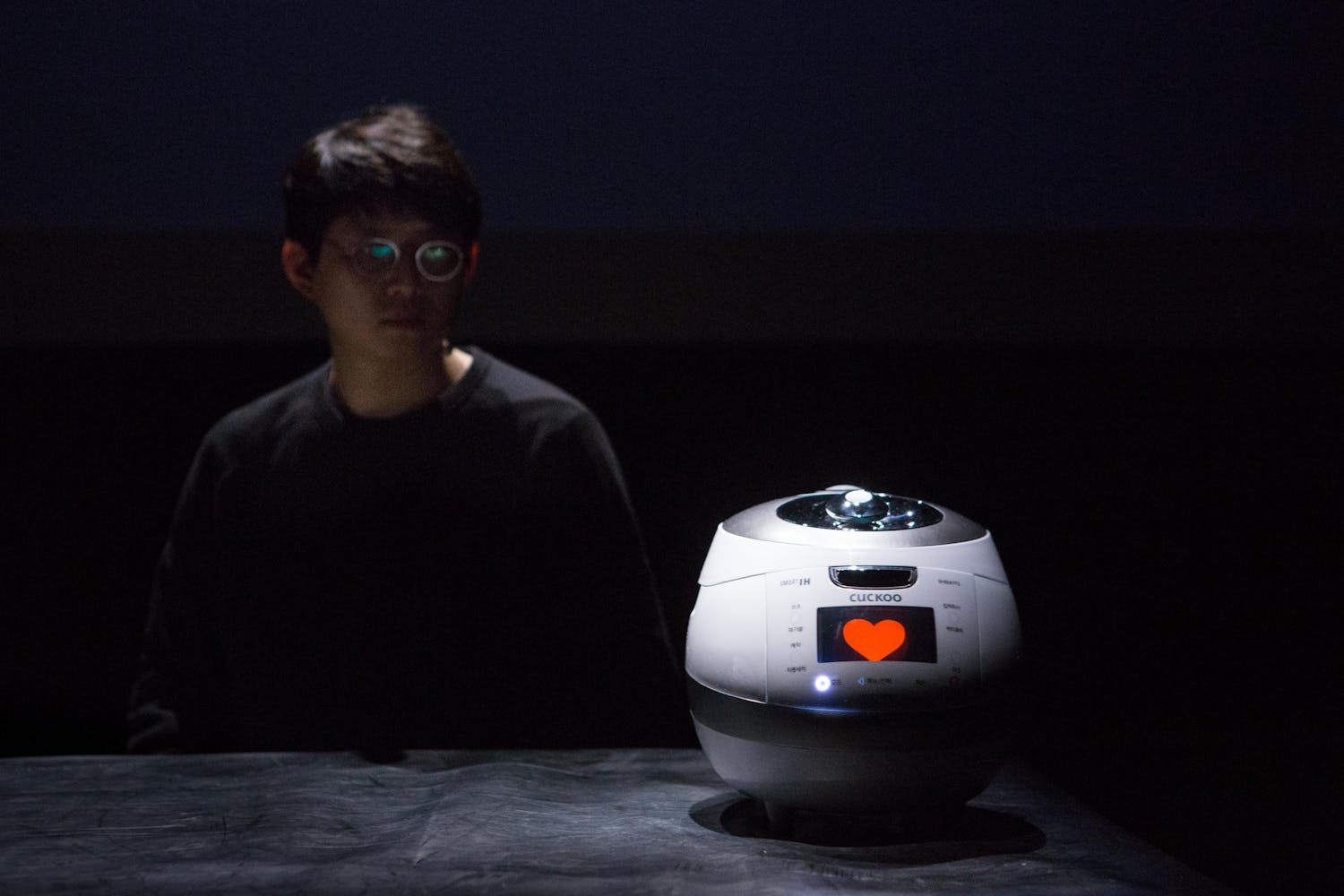 A performer with medium skin sits next to a rice cooker with a heart on its screen.