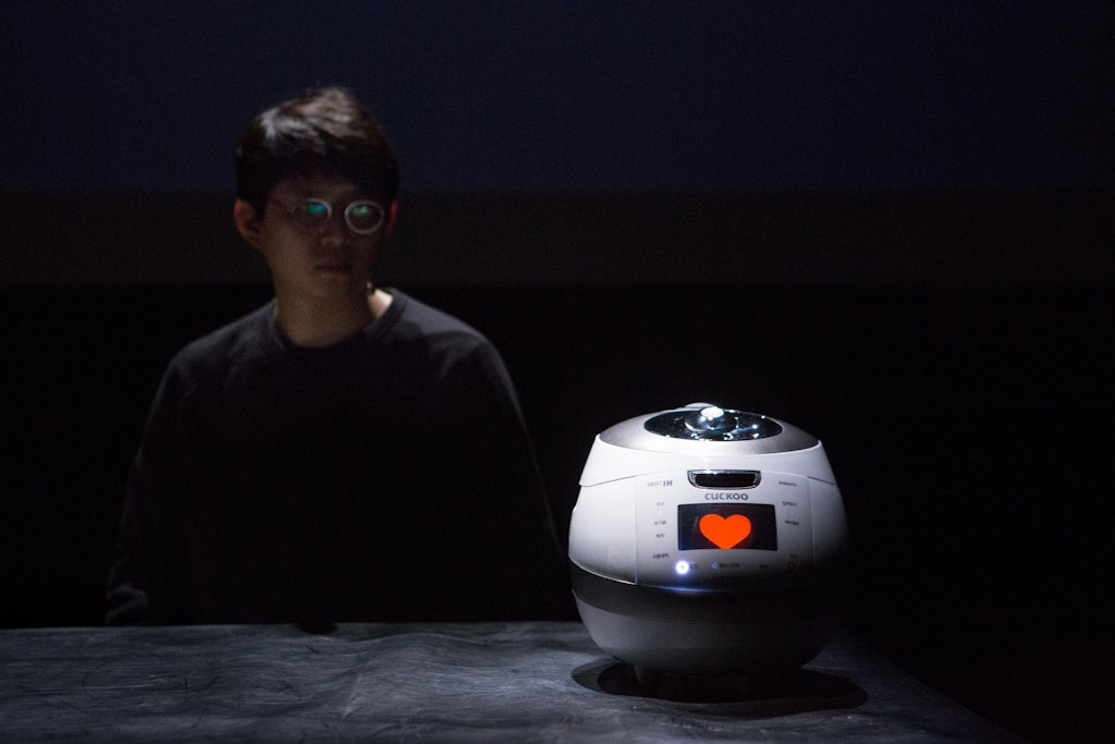 A performer with medium skin sits next to a rice cooker with a heart on its screen.