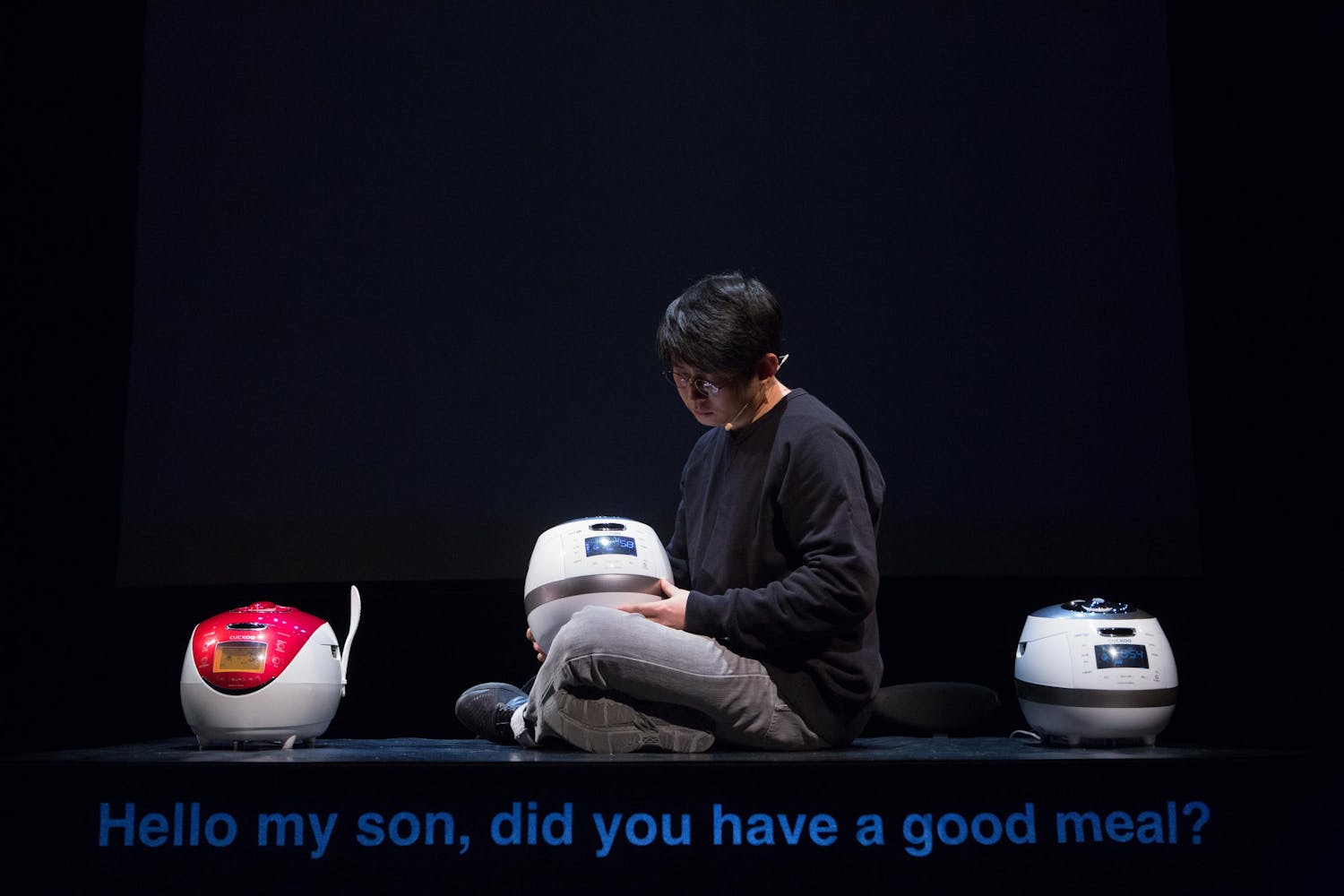 A performer with medium skin sits with a rice cooker in their lap. The caption reads 