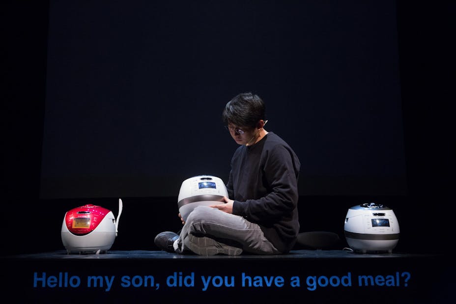A performer with medium skin sits with a rice cooker in their lap. The caption reads "Hello my son, did you have a good meal?".
