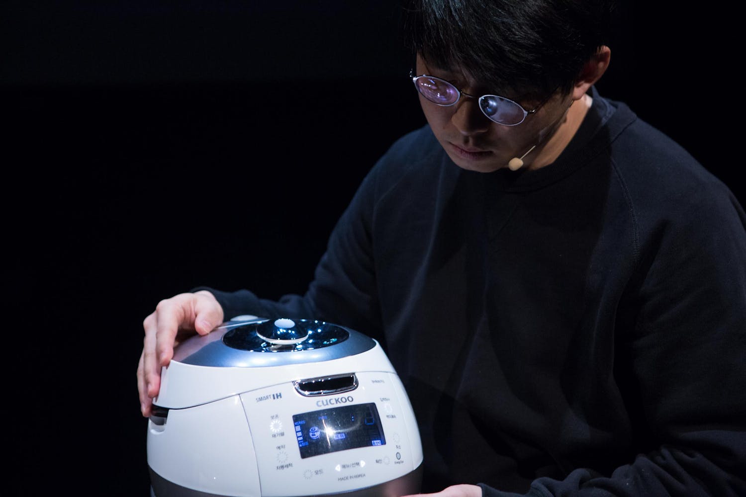 A performer with medium skin holds a rice cooker on his lap.