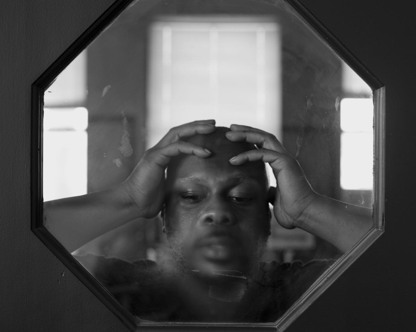 Black and white photo of a man with his hands on his head through an octagonal window that’s fogged in front of his mouth.