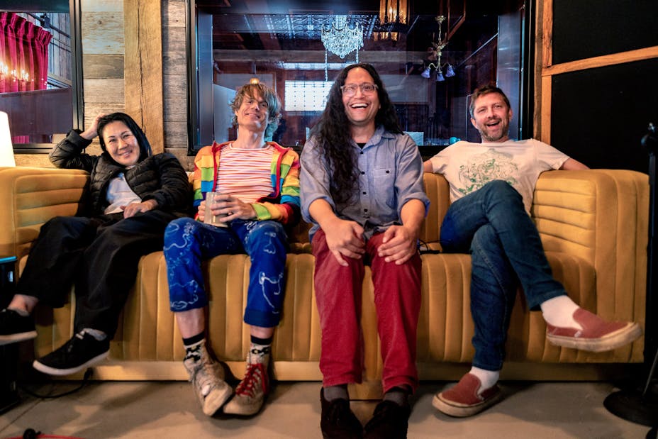 Four musicians sit on a yellow couch with eclectic clothing.