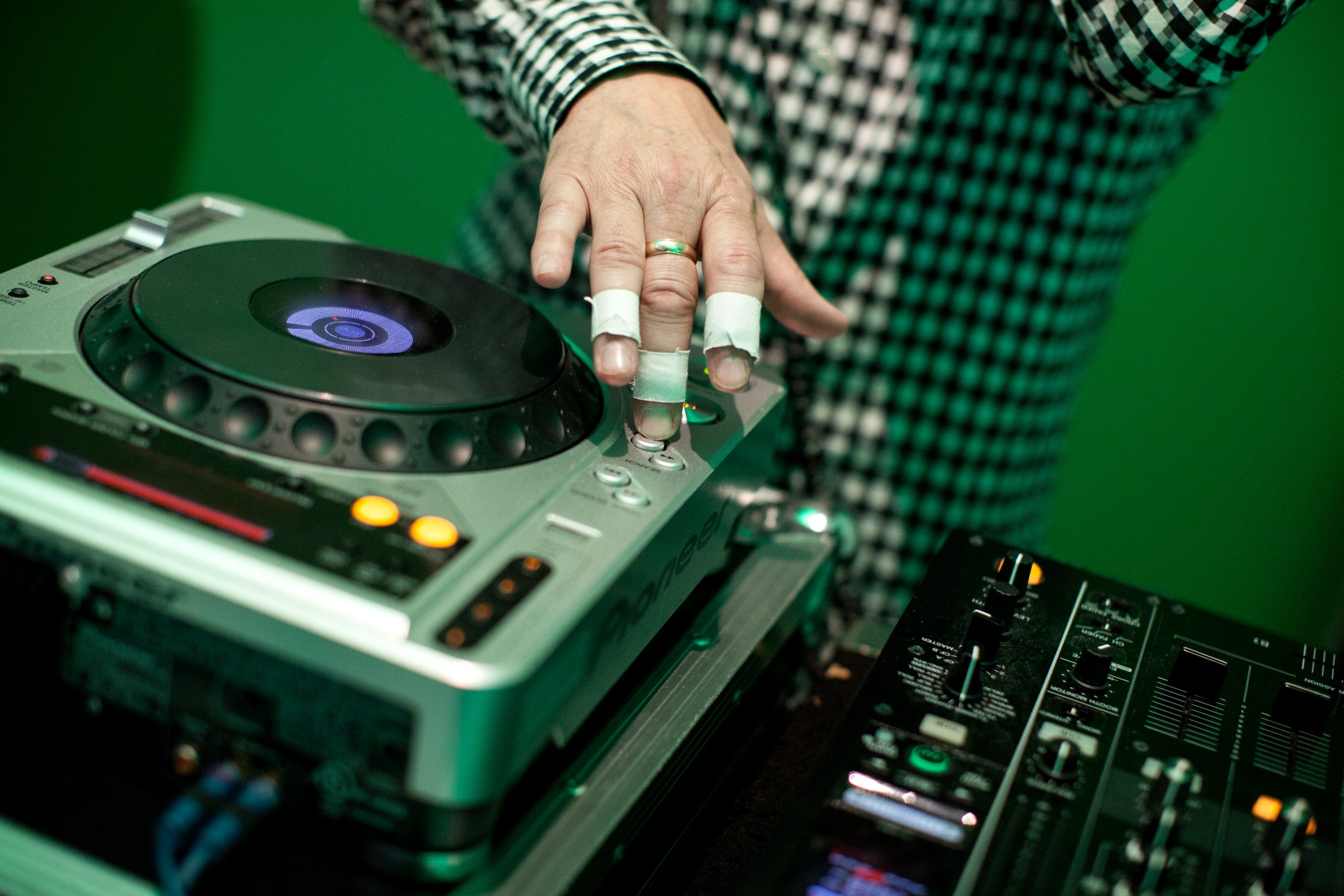 A DJ with taped fingers adjusts equipment, wearing a black and white checkered shirt against a green background.