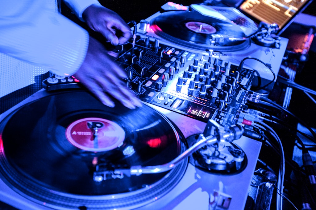 A DJs hand spinning vinyl records on turntables with a mixer, illuminated by blue stage lights.