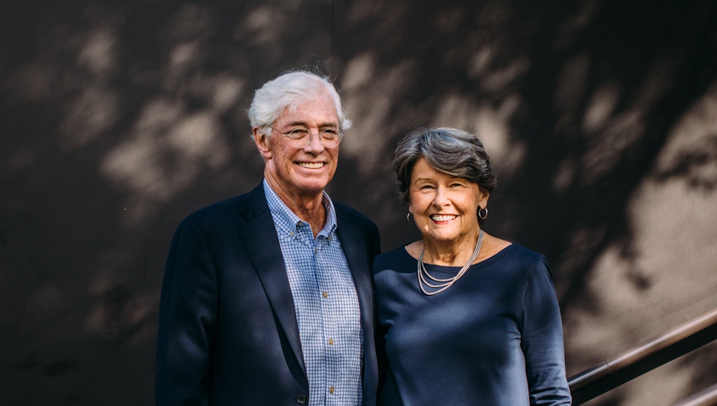 An older couple stand outside against a concrete wall and smile at the viewer.