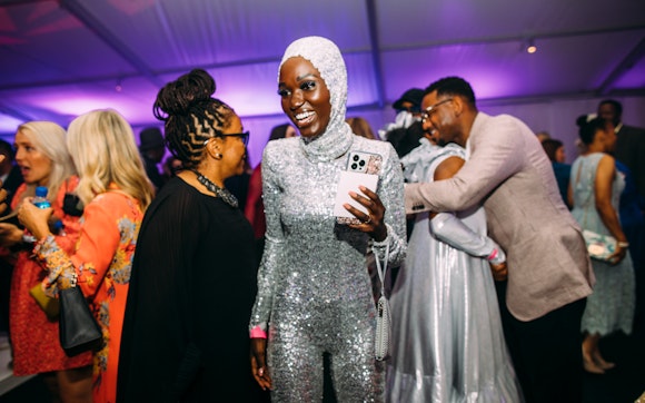 A woman in a full body silver sequin jumpsuit smiles while walking through the dancefloor at a festive party.