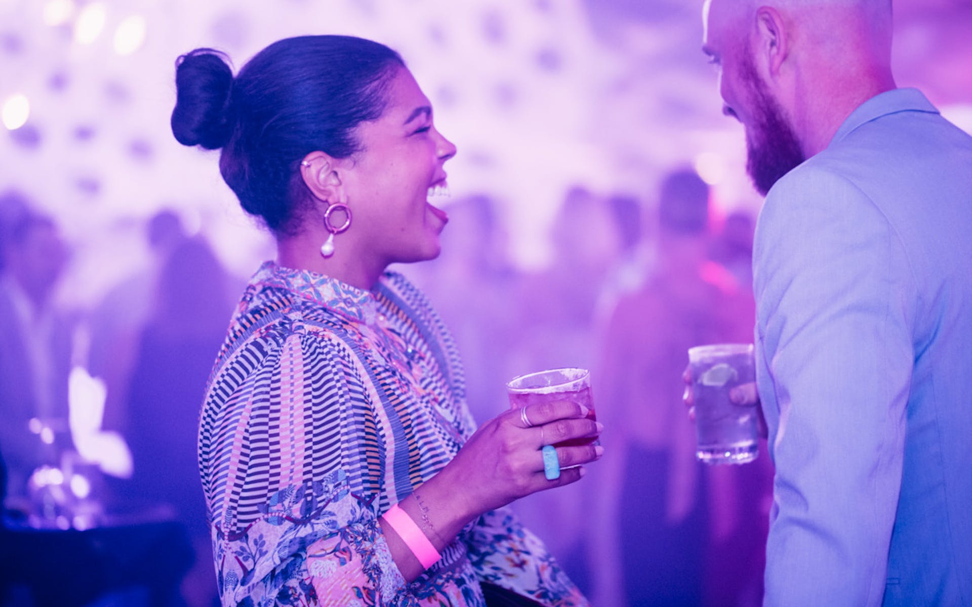 A woman laughs while holding a drink and talking to a man at a party where the room is bathed in purple light.