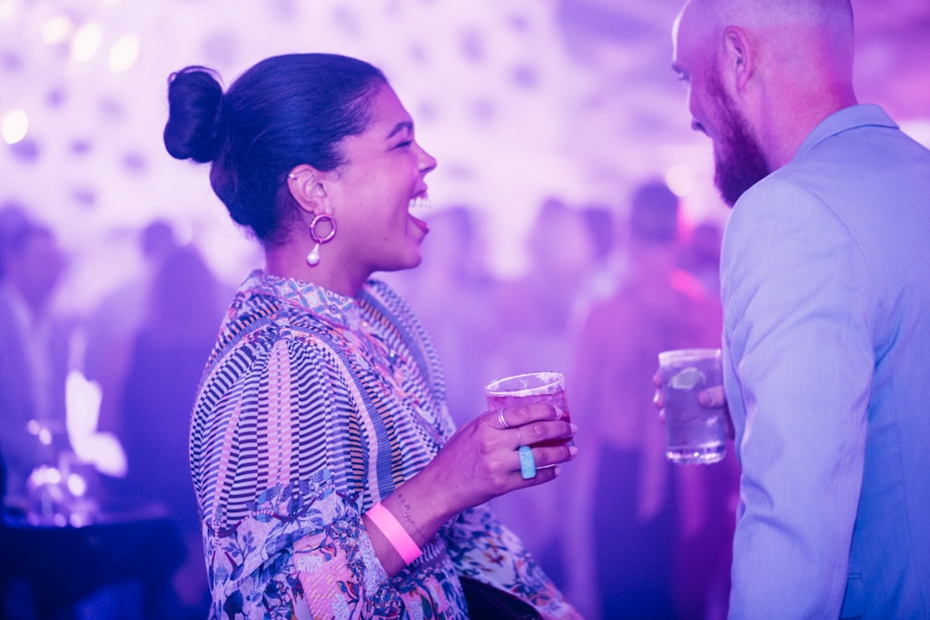 A woman laughs while holding a drink and talking to a man at a party where the room is bathed in purple light.