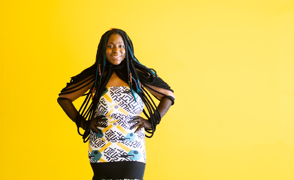 A woman stands in front of a yellow wall with her hands on her hips and smiles at the viewer.