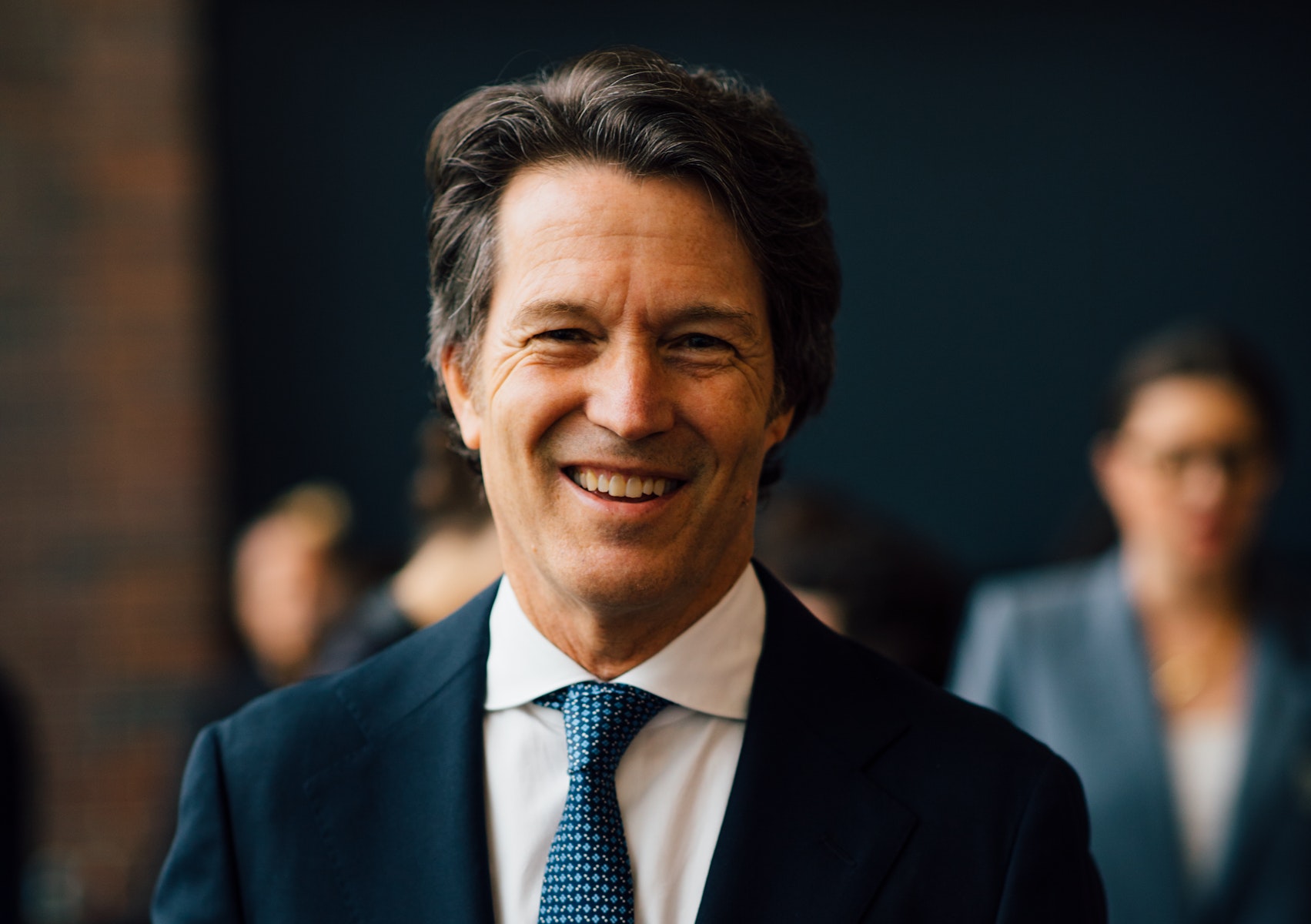 A man with light skin smiles at the viewer while wearing a dark suit coat, white shirt, and tie.
