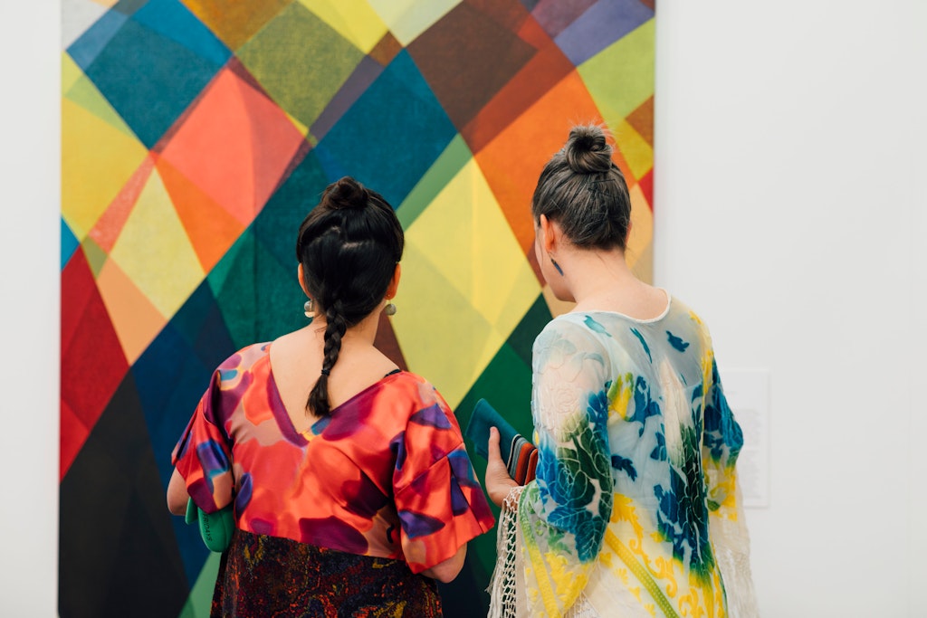 Two people stand in front of a large white wall with a colorful geometric painting resembling a warped multicolored checkerboard in pinks, yellows, oranges, greens, and blues.