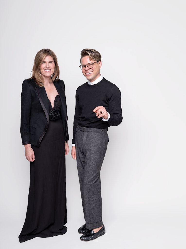 Two adults with light skin wearing formal clothing stand in front of a white background and smile at the viewer