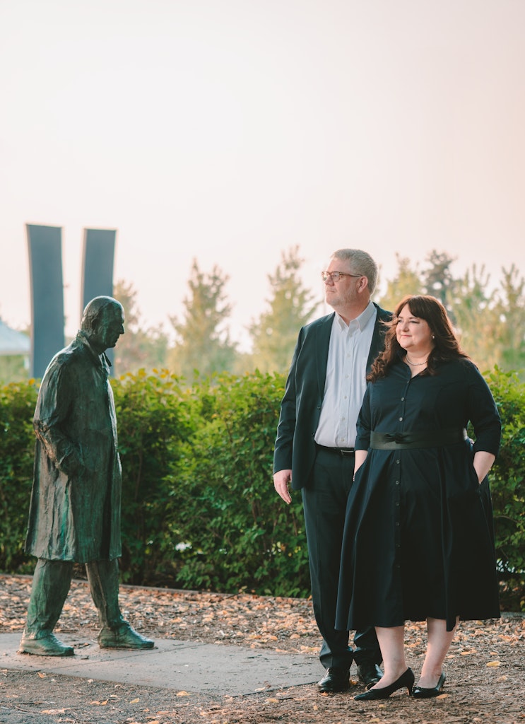 An adult couple with light skin and dark clothes stands in a garden outside looking at a statue