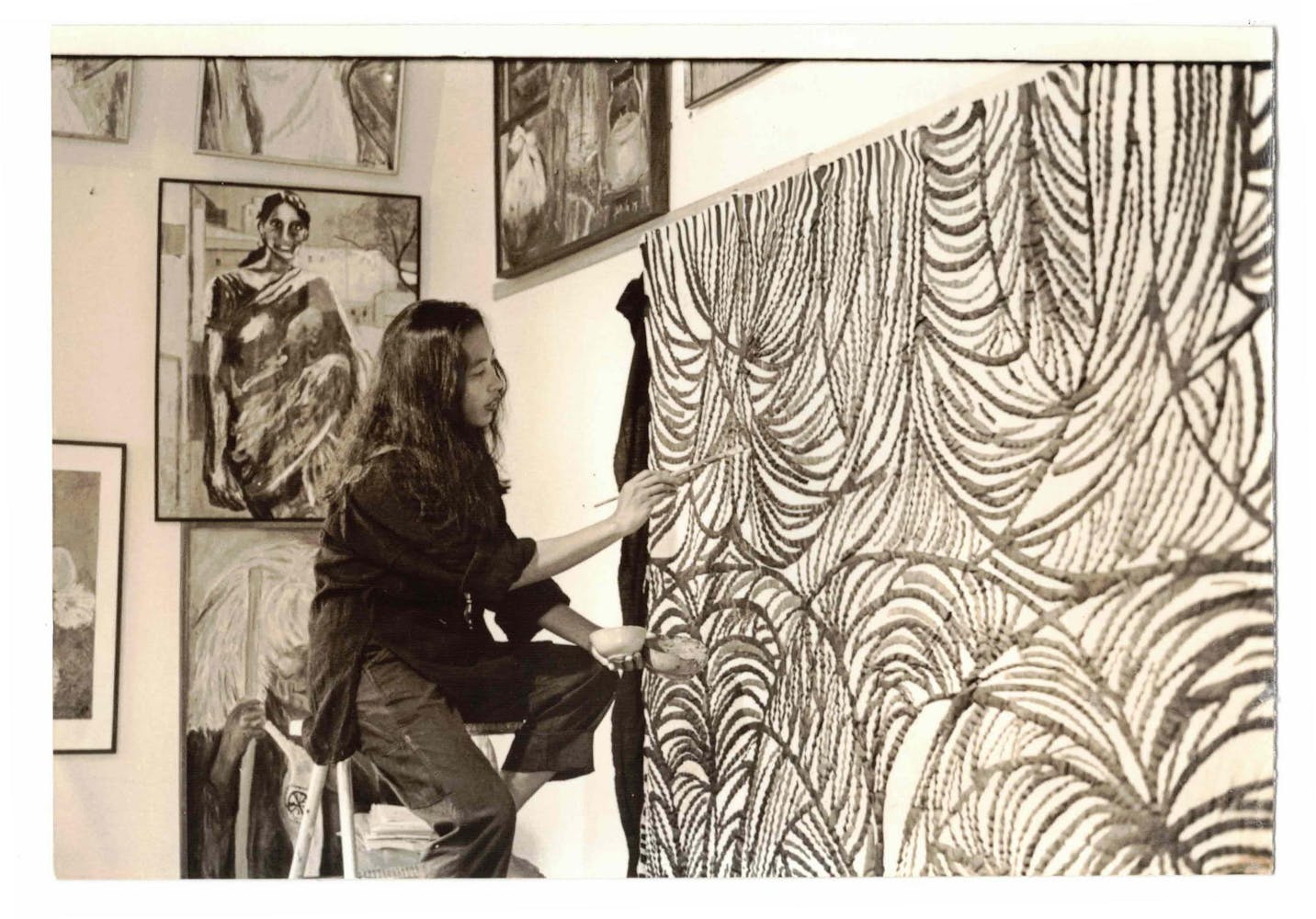 A woman sits on a ladder while working on a large textile painting in their studio.