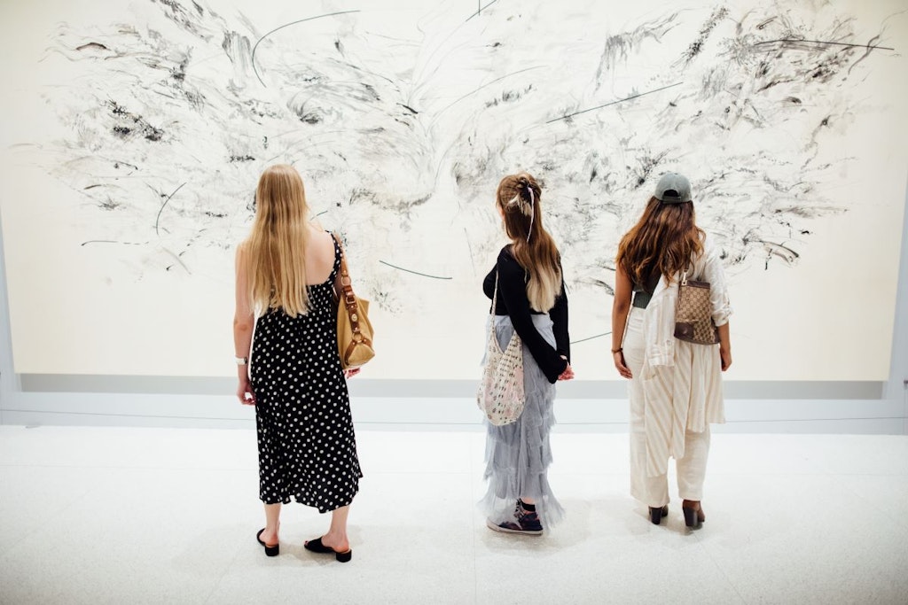 Three adults face away from the viewer and look at a large piece of black and white art in a gallery