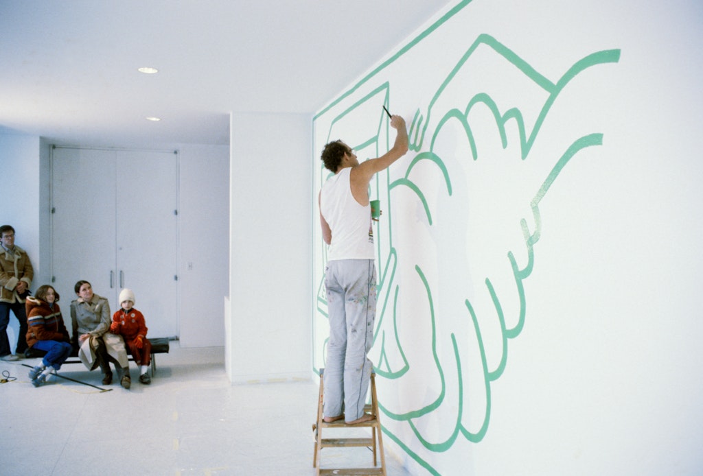 An adult with light skin and paint-covered pants, white tank top, stands barefoot, painting a mural on a white wall.