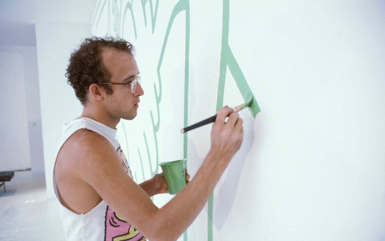 ecp1984KH_concourse_010 A light skinned man with glasses in a white sleeveless shirt paints on a wall with green paint.